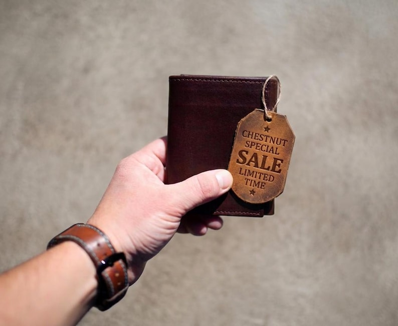 May include: A dark brown leather wallet with a matching leather tag. The tag reads "CHESTNUT SPECIAL SALE LIMITED TIME" and is attached with a string. The wallet is held in a hand, with a brown leather watch visible.