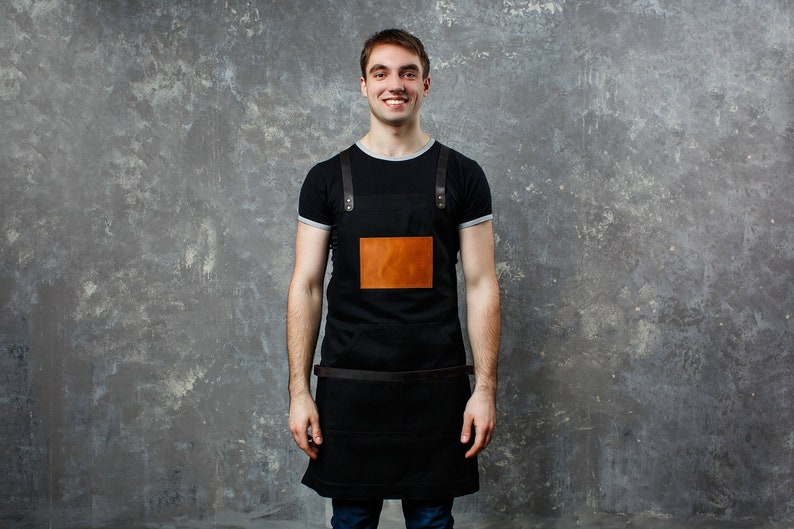 May include: A man wearing a black apron with a brown leather patch and a black t-shirt. He is standing in front of a gray textured wall.