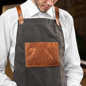 May include: A brown canvas apron with leather straps and a brown leather patch pocket. The apron is worn by a person with a white shirt.