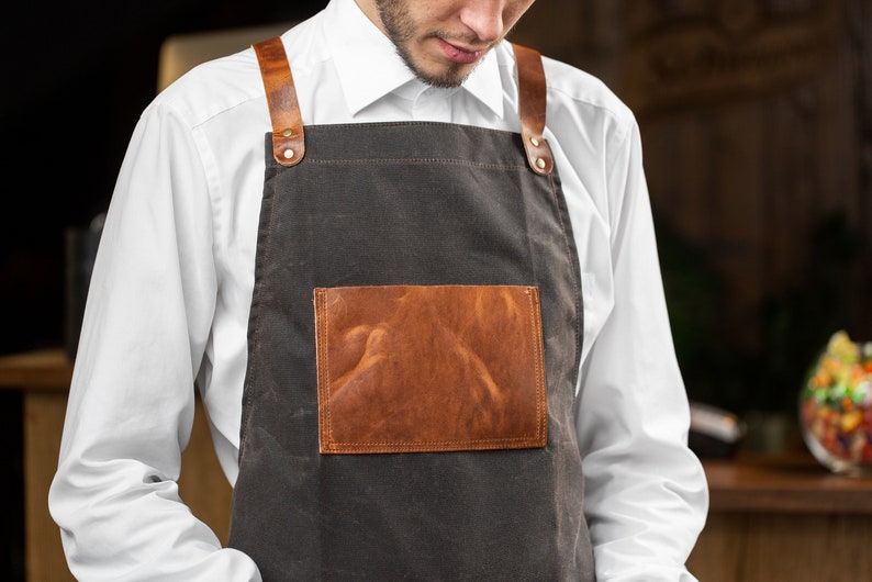 May include: A brown canvas apron with leather straps and a brown leather patch pocket. The apron is worn by a person with a white shirt.
