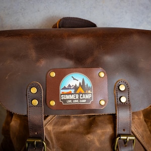 Puede incluir: Una mochila de cuero marr&oacute;n con cierre de solapa y herrajes de lat&oacute;n. La parte delantera presenta un parche rectangular con un gr&aacute;fico de "Summer Camp", que incluye una monta&ntilde;a, un lago y una escena de tienda de campa&ntilde;a. Tambi&eacute;n se incluye el texto "Live, Love, Camp".