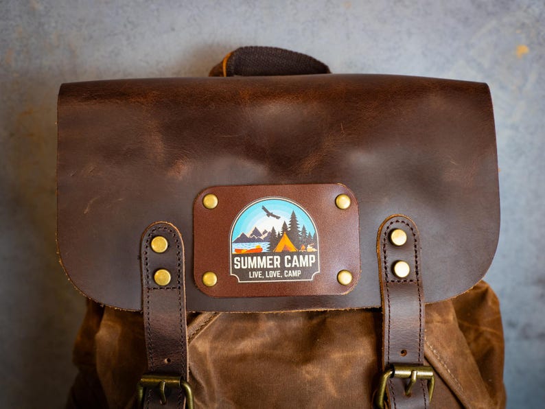 Puede incluir: Una mochila de cuero marr&oacute;n con cierre de solapa y herrajes de lat&oacute;n. La parte delantera presenta un parche rectangular con un gr&aacute;fico de "Summer Camp", que incluye una monta&ntilde;a, un lago y una escena de tienda de campa&ntilde;a. Tambi&eacute;n se incluye el texto "Live, Love, Camp".