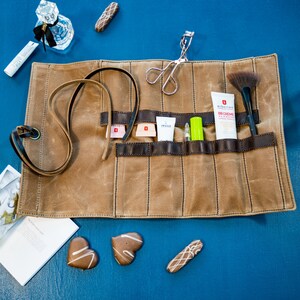 May include: A brown canvas roll-up makeup bag with multiple compartments. The bag is unrolled and contains various makeup and beauty products, including a lash curler, a brush, a tube of BB cream, a small bottle of lotion, and a green tube of lip balm. The bag is on a blue surface with various chocolate candies.
