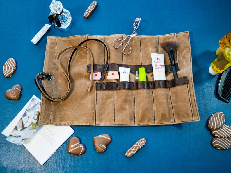 May include: A brown canvas roll-up makeup bag with multiple compartments. The bag is unrolled and contains various makeup and beauty products, including a lash curler, a brush, a tube of BB cream, a small bottle of lotion, and a green tube of lip balm. The bag is on a blue surface with various chocolate candies.