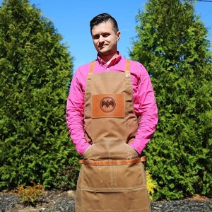 May include: A brown canvas apron with a leather patch featuring a bat logo. The apron has two large pockets and adjustable straps. The person wearing the apron is wearing a pink shirt.