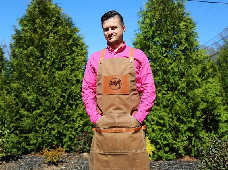 May include: A brown canvas apron with a leather patch featuring a bat logo. The apron has two large pockets and adjustable straps. The person wearing the apron is wearing a pink shirt.