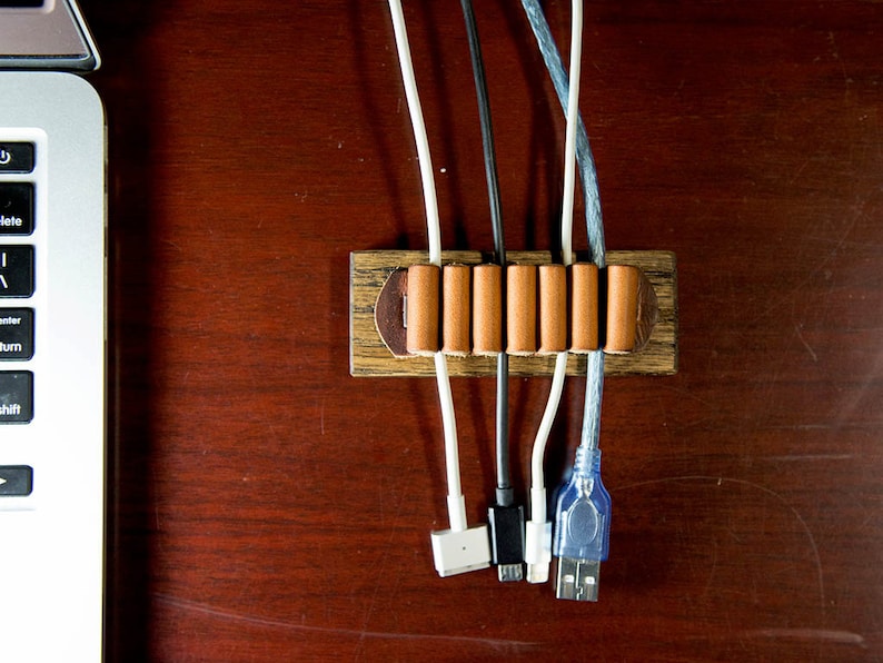 Puede incluir: Un organizador de cables de madera con seis correas de cuero marr&oacute;n para sujetar los cables en su lugar. El organizador est&aacute; sobre una superficie de madera marr&oacute;n con varios cables que pasan a trav&eacute;s de &eacute;l.