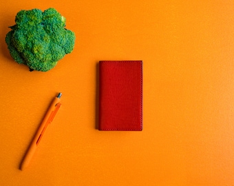 May include: A red leather wallet on a bright orange background with a green broccoli and an orange pen.