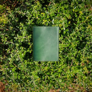 May include: A green leather journal with stitched edges sits on a bed of green foliage. The journal's cover is a solid, rich green colour, and the stitching provides a subtle decorative detail. The background is a dense arrangement of green leaves and branches.