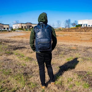 May include: A person wearing a black hooded sweatshirt and black jeans is standing in a field with a black leather backpack on their back. The person is looking at a building in the distance.