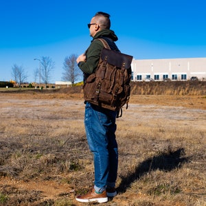 Puede incluir: Una mochila de lona y cuero marr&oacute;n con m&uacute;ltiples bolsillos y correas. La mochila tiene un cierre de solapa y un asa superior. La bolsa la lleva una persona con vaqueros azules y una sudadera con capucha verde, de pie en un campo en un d&iacute;a soleado.