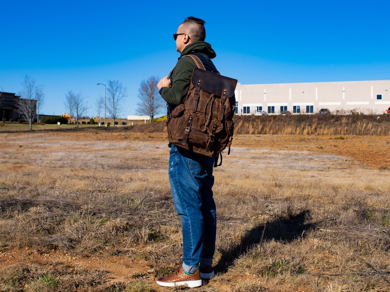 Puede incluir: Una mochila de lona y cuero marr&oacute;n con m&uacute;ltiples bolsillos y correas. La mochila tiene un cierre de solapa y un asa superior. La bolsa la lleva una persona con vaqueros azules y una sudadera con capucha verde, de pie en un campo en un d&iacute;a soleado.
