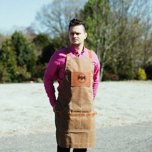 May include: A man wearing a pink button-down shirt and a brown canvas apron with a leather patch featuring a bat logo. The apron has adjustable straps and multiple pockets.