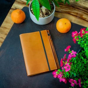 May include: A brown leather notebook with a black elastic closure and a black pen on a dark brown desk with a potted plant and two oranges.