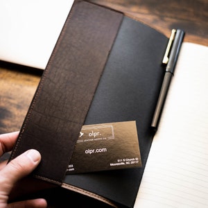 K&ouml;nnte beinhalten: Ein braunes Lederjournal mit einem schwarzen Stift und einer Visitenkarte mit der Aufschrift "olpr. LEATHER GOODS CO olpr.com 511 S Church St Mooresville, NC 28117"