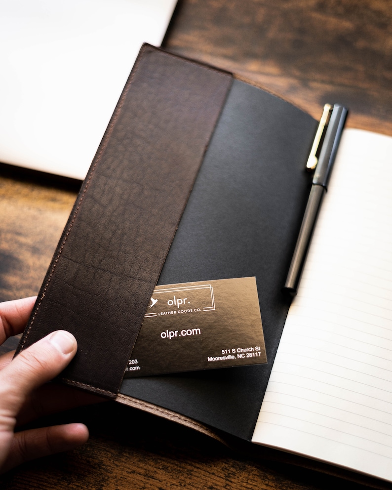 K&ouml;nnte beinhalten: Ein braunes Lederjournal mit einem schwarzen Stift und einer Visitenkarte mit der Aufschrift "olpr. LEATHER GOODS CO olpr.com 511 S Church St Mooresville, NC 28117"
