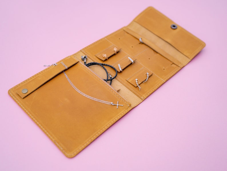 May include: Open, tan leather jewelry organizer with various compartments and snap closure. The organizer holds a silver cross necklace, earrings, and a black braided bracelet. The background is a solid pink color.