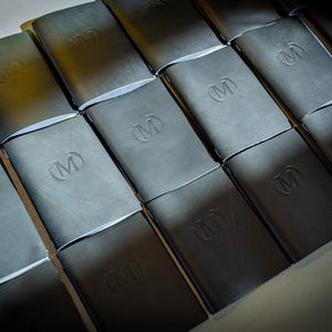 May include: A collection of rectangular black notebooks arranged in a grid pattern. Each notebook features a smooth, matte cover with a debossed letter M logo centered on the front. The notebooks are positioned side by side in rows.