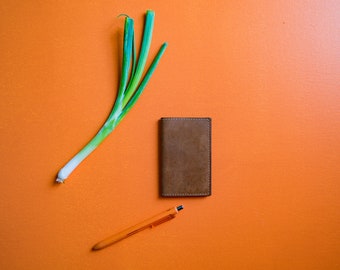 Könnte beinhalten: Eine braune Ledergeldbörse, eine grüne Zwiebel und ein orangefarbener Stift auf einem leuchtend orangenen Hintergrund.