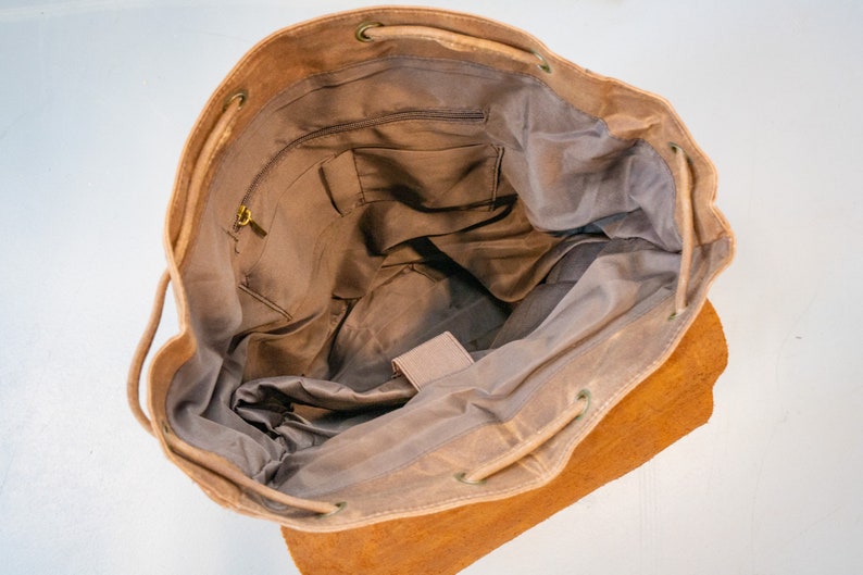 Puede incluir: Vista abierta de una mochila de lona color beige con cierre de cord&oacute;n. El interior presenta m&uacute;ltiples bolsillos y un peque&ntilde;o compartimento con cremallera. La bolsa tiene una base de cuero y herrajes color lat&oacute;n.