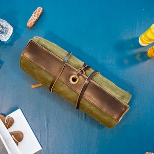 K&ouml;nnte beinhalten: Eine gr&uuml;ne und braune Canvas-Rolltasche mit einem Lederriemen und einer Messing-Schnalle. Die Tasche ist aufgerollt und mit dem Riemen gebunden.