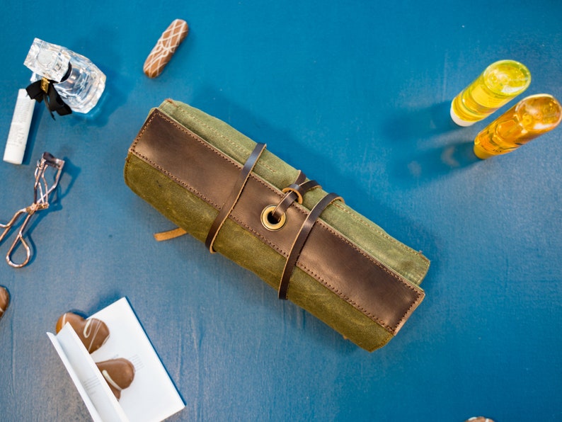 K&ouml;nnte beinhalten: Eine gr&uuml;ne und braune Canvas-Rolltasche mit einem Lederriemen und einer Messing-Schnalle. Die Tasche ist aufgerollt und mit dem Riemen gebunden.