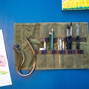 K&ouml;nnte beinhalten: Eine gr&uuml;ne Canvas-Rolltasche mit braunen Lederriemen und einem Ledertragegurt. Die Tasche ist ge&ouml;ffnet und enth&auml;lt eine Vielzahl von Kunstbedarfsartikeln, darunter Bleistifte, Stifte und Pinsel.
