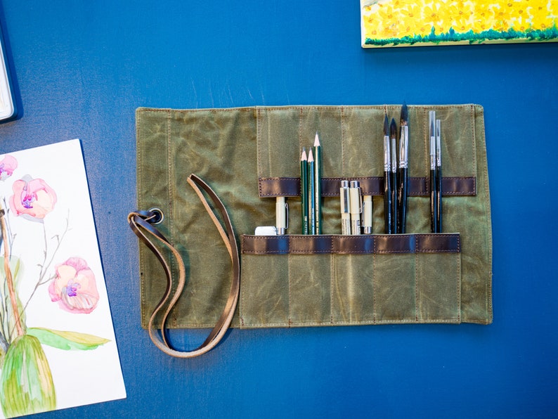 K&ouml;nnte beinhalten: Eine gr&uuml;ne Canvas-Rolltasche mit braunen Lederriemen und einem Ledertragegurt. Die Tasche ist ge&ouml;ffnet und enth&auml;lt eine Vielzahl von Kunstbedarfsartikeln, darunter Bleistifte, Stifte und Pinsel.