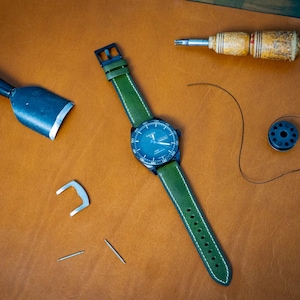 Puede incluir: Un reloj de pulsera con correa de cuero verde y esfera azul oscuro, junto con herramientas de marroquiner&iacute;a. El reloj tiene costuras blancas en la correa. Las herramientas incluyen un cuchillo de cuero, un destornillador, agujas y una bobina de hilo. Los art&iacute;culos est&aacute;n sobre una superficie de cuero marr&oacute;n.