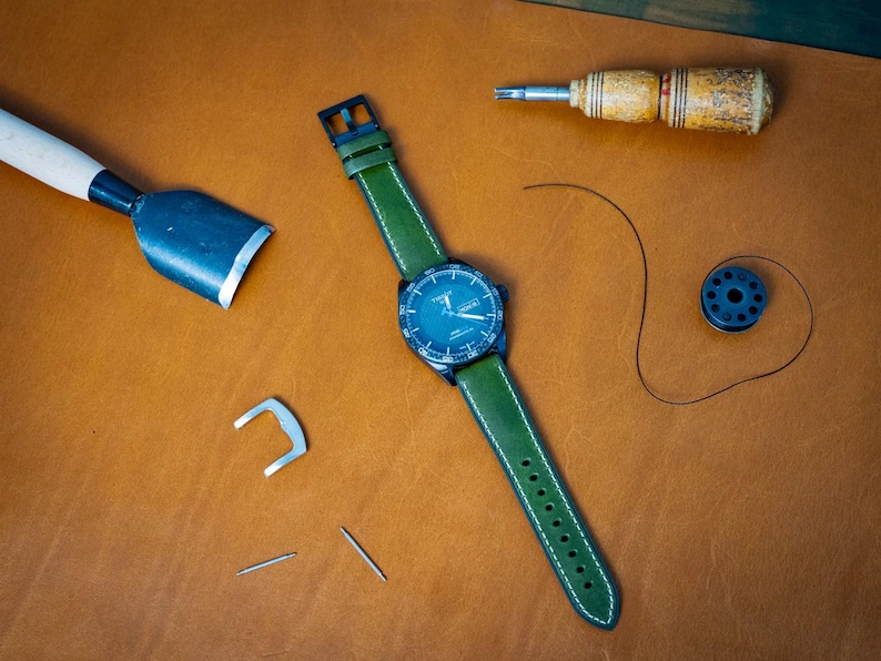 Puede incluir: Un reloj de pulsera con correa de cuero verde y esfera azul oscuro, junto con herramientas de marroquiner&iacute;a. El reloj tiene costuras blancas en la correa. Las herramientas incluyen un cuchillo de cuero, un destornillador, agujas y una bobina de hilo. Los art&iacute;culos est&aacute;n sobre una superficie de cuero marr&oacute;n.