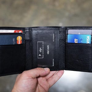 May include: Black leather trifold wallet, open to display card slots and a clear ID window. The wallet holds a debit card, a Mastercard, and a Visa card. The brand name "olpr" is visible.