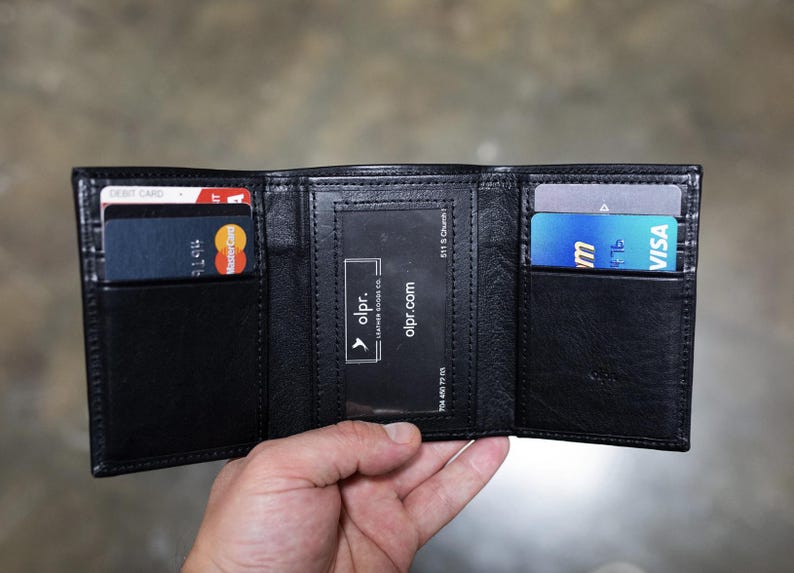 May include: Black leather trifold wallet, open to display card slots and a clear ID window. The wallet holds a debit card, a Mastercard, and a Visa card. The brand name "olpr" is visible.