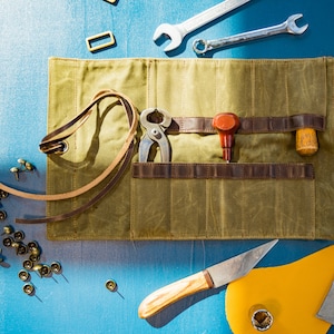 K&ouml;nnte beinhalten: Eine gr&uuml;ne Canvas-Rolltasche mit braunen Lederriemen und Taschen. Die Tasche ist offen und enth&auml;lt verschiedene Werkzeuge, darunter eine Zange, einen kleinen Hammer, einen Schraubendreher und einen Lederstanzer. Au&szlig;erdem liegen einige Metallnieten und Lederstreifen um die Tasche herum.