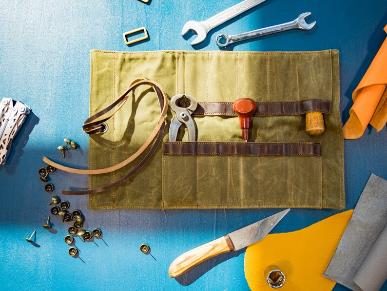 K&ouml;nnte beinhalten: Eine gr&uuml;ne Canvas-Rolltasche mit braunen Lederriemen und Taschen. Die Tasche ist offen und enth&auml;lt verschiedene Werkzeuge, darunter eine Zange, einen kleinen Hammer, einen Schraubendreher und einen Lederstanzer. Au&szlig;erdem liegen einige Metallnieten und Lederstreifen um die Tasche herum.