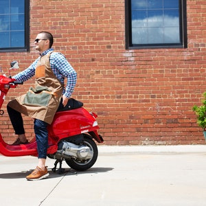 May include: A person wearing a brown leather and canvas apron is riding a red Vespa scooter in front of a brick wall. There is a sign that reads "No Parking Any Time" on the wall.
