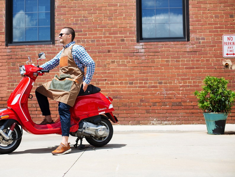 May include: A person wearing a brown leather and canvas apron is riding a red Vespa scooter in front of a brick wall. There is a sign that reads "No Parking Any Time" on the wall.