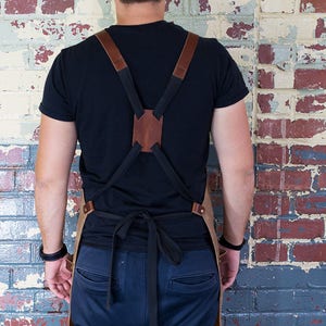 May include: A brown leather apron with adjustable straps and a black canvas bib. The apron is worn by a person standing in front of a brick wall.