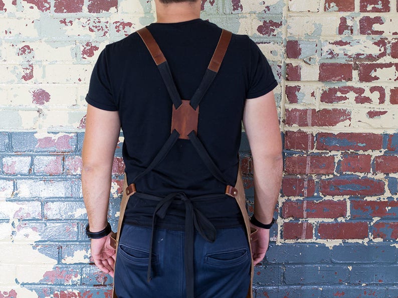 May include: A brown leather apron with adjustable straps and a black canvas bib. The apron is worn by a person standing in front of a brick wall.