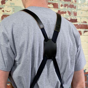 May include: A person wearing a gray t-shirt with black suspenders. The suspenders have a leather strap and a black buckle.