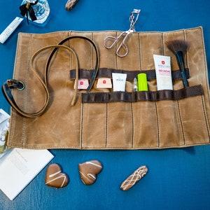 May include: A brown canvas roll-up pouch with multiple pockets for storing makeup and beauty supplies. The pouch is unrolled and shows a variety of items, including a makeup brush, a tube of face cream, a lip gloss, and a pair of tweezers.