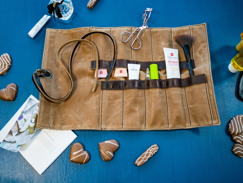 May include: A brown canvas roll-up pouch with multiple pockets for storing makeup and beauty supplies. The pouch is unrolled and shows a variety of items, including a makeup brush, a tube of face cream, a lip gloss, and a pair of tweezers.