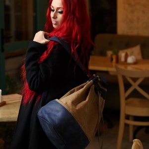 May include: A woman with long, bright red hair is wearing a black coat and a blue and brown drawstring backpack. The backpack has a large, round shape and is worn over one shoulder.