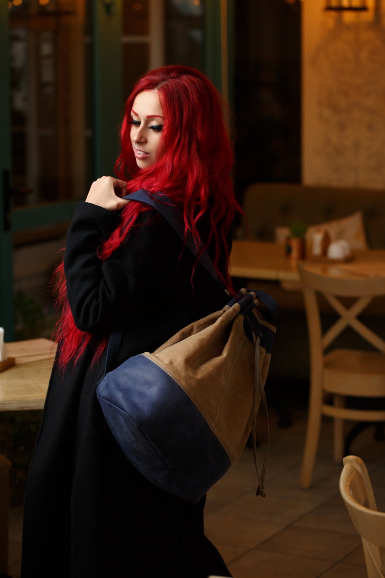 May include: A woman with long, bright red hair is wearing a black coat and a blue and brown drawstring backpack. The backpack has a large, round shape and is worn over one shoulder.