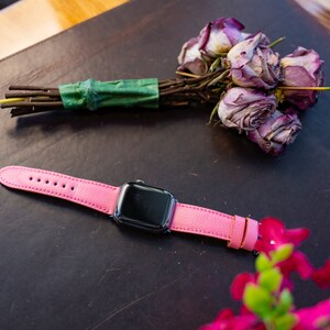 Puede incluir: Una correa de reloj de cuero rosa con una esfera de Apple Watch negra. El reloj est&aacute; sobre una superficie de cuero marr&oacute;n con un ramo de rosas secas en el fondo.