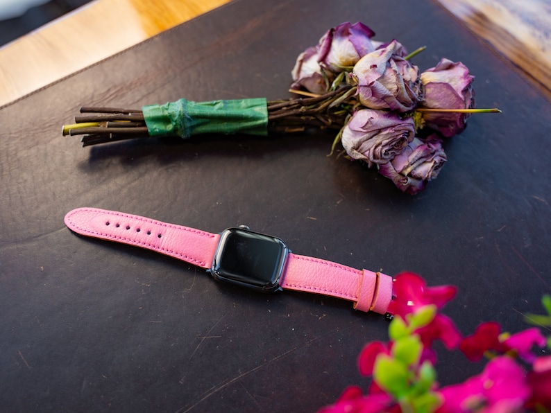 Puede incluir: Una correa de reloj de cuero rosa con una esfera de Apple Watch negra. El reloj est&aacute; sobre una superficie de cuero marr&oacute;n con un ramo de rosas secas en el fondo.