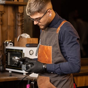 May include: A man wearing a brown leather and canvas apron with a black shirt and black gloves is holding a black and silver electric razor.