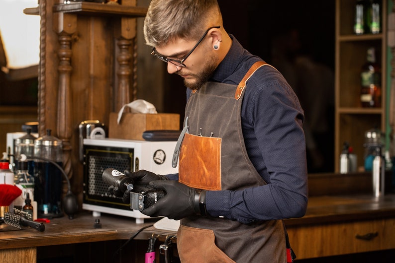 May include: A man wearing a brown leather and canvas apron with a black shirt and black gloves is holding a black and silver electric razor.