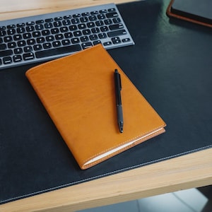 May include: A leather-bound journal in a warm tan hue rests on a black desk mat, accompanied by a black pen. A silver keyboard is positioned above the journal. The scene suggests a workspace or office setting, ideal for note-taking or journaling.
