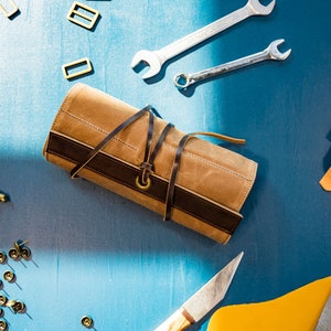May include: A brown leather roll-up case with a dark brown leather strap and a brass buckle. The case is on a blue surface with various tools and leather scraps.