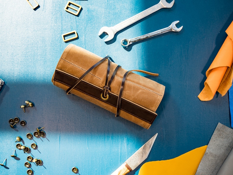 May include: A brown leather roll-up case with a dark brown leather strap and a brass buckle. The case is on a blue surface with various tools and leather scraps.